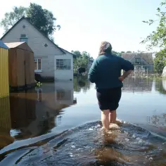 Kobieta brodzi w wodzie po powodzi, która zalała domy i podwórka. W tle widać zalane budynki i drzewa.