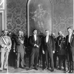 Uroczyste spotkanie panów w galowych strojach, w tym wojskowych, przed obrazem. W tle bogata tapeta. bbwr.