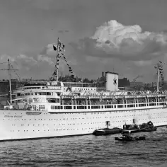 Ogromny statek pasażerski Wilhelm Gustloff cumuje w porcie, otoczony mniejszymi łodziami. W tle widoczne miasto z wieżami kościołów.