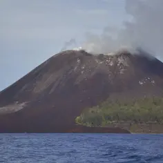 Aktywny wulkan Krakatau dymi, jego zbocza pokryte są ciemną lawą, a u podnóża rośnie bujna roślinność.