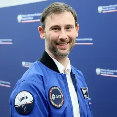 Uśmiechnięty mężczyzna w niebieskiej kurtce z naszywkami ESA i Ignis, jak prawdziwi astronauci, pozuje na tle niebieskiej ściany z logo ministerstwa.