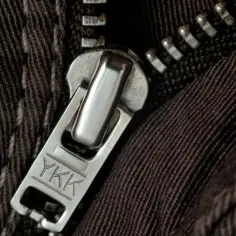 Zbliżenie na metalowy suwak YKK na brązowych spodniach. Kiedy wynaleziono suwak, zmienił sposób zapinania ubrań.