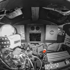 Wnętrze statku kosmicznego, widok z perspektywy astronauty. Widać hełm, panele sterowania z logo NASA i napisem "CALLISTO TECH DEMO". Mała, czerwona maskotka pingwina symbolizuje ducha misji Apollo 13.