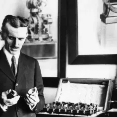 Nikola Tesla w garniturze trzymający elementy aparatury elektrycznej, obok skrzynki z urządzeniami.