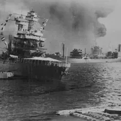 Dym unosi się nad zatoką po ataku na Pearl Harbor. Okręt wojenny z banderami w tle.