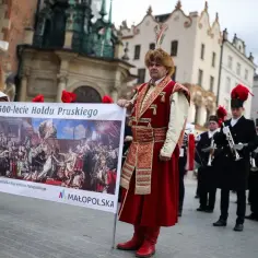Mężczyzna w stroju historycznym trzyma baner z obrazem przedstawiającym hołd pruski.
