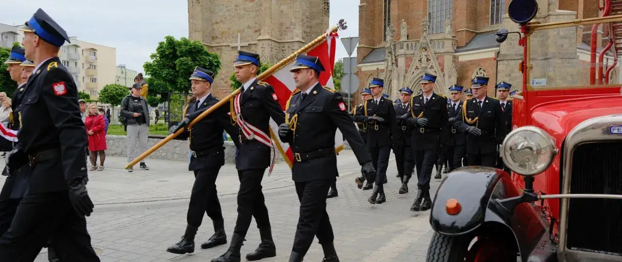 Uroczysty przemarsz strażaków w mundurach z biało-czerwoną flagą. Obchody 4 maja w mieście.