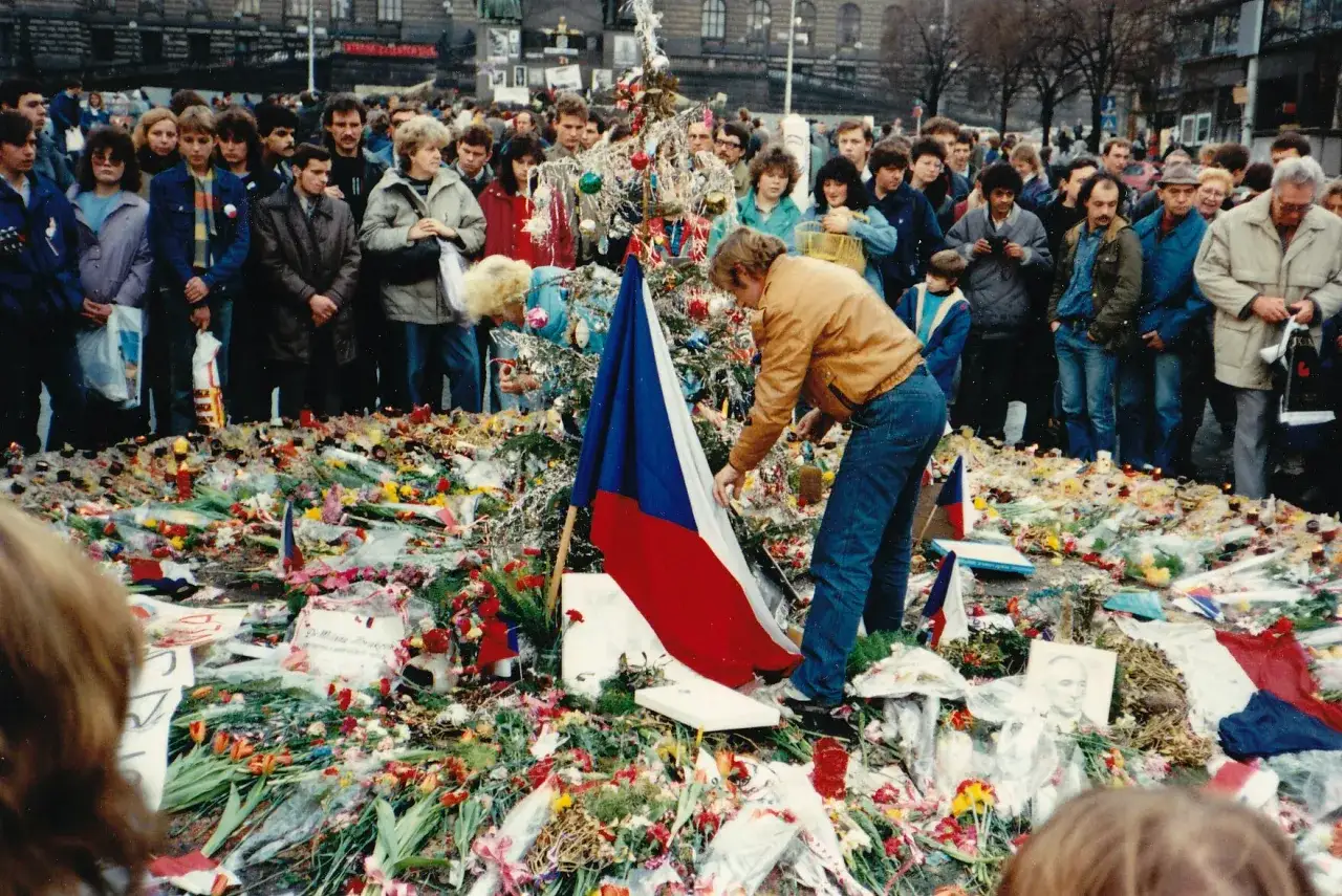 Tłum ludzi składa kwiaty i zapala znicze pod choinką, symbolizując upadek komunizmu. Flaga Czechosłowacji jest widoczna.