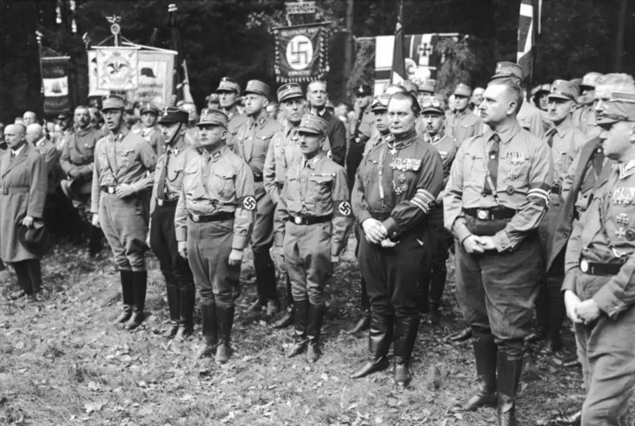 Grupa mężczyzn w brunatnych koszulach i czapkach stoi na tle sztandarów.